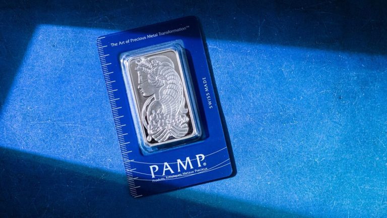 Silver bar in PAMP packaging on a textured blue surface with sunlight.